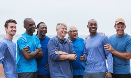 Multi-ethnic group of men standing together, smiling, supporting one another. Mixed ages ranging from 20s to 70s.